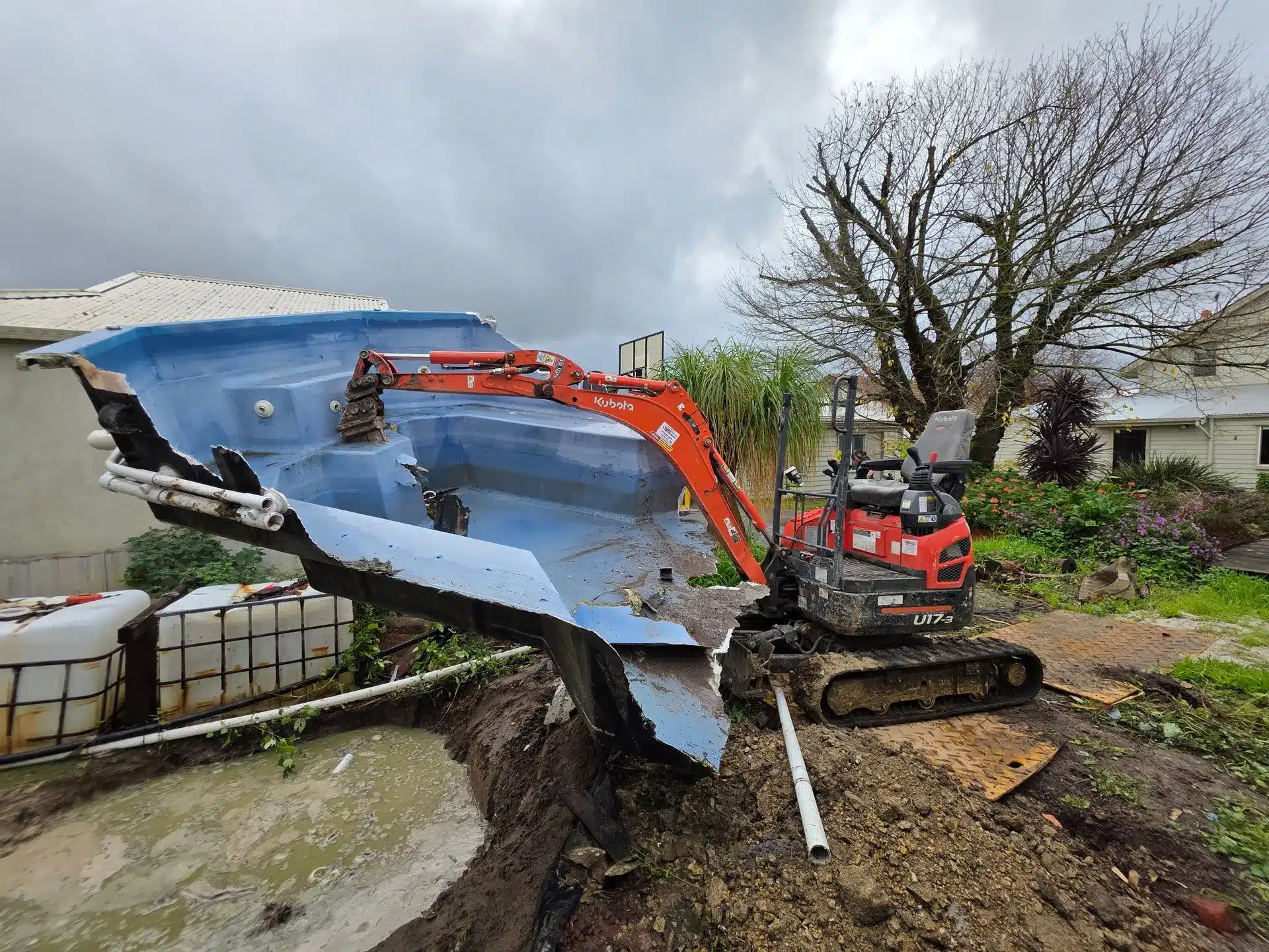Excavator lifting a blue fibreglass pool shell out of the ground